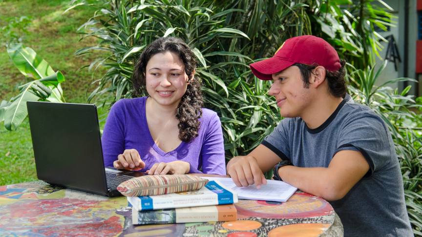 Zwei Studierende sitzen mit einem Laptop und Büchern an einem Tisch im Garten.